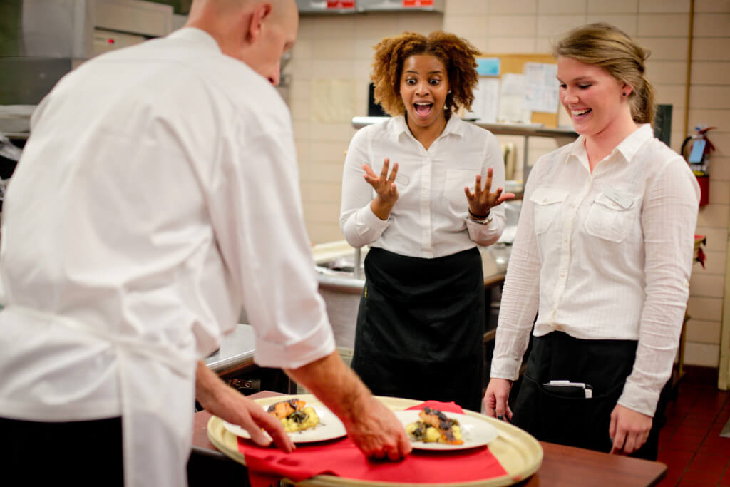 Photo by Michelle LaVigne/Creative Services Senior Ahley Hahn (right,) in nutrition and dietetics from Geneva, Ill., listens as junior Denise Worrell in nutrition and dietetics from Chicago, Ill., describes a moment she had while serving as Chef Bryan Flower gives the servers a lesson in how to present the meal on a tray.